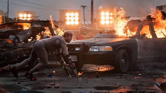 A person in a grey bodysuit runs past burning vehicles under bright lights at dusk.