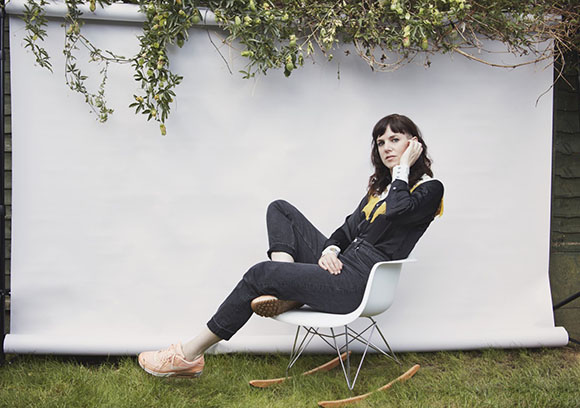 A woman in black clothing and pink sneakers sits in a white chair against a white backdrop in a grassy area.