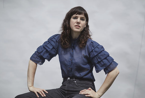 A woman with dark hair and bangs wearing a navy blue ruffled blouse and black jeans, posing against a white wall.