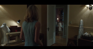 A woman in a light blue sleeveless top stands in a room looking toward an open doorway.