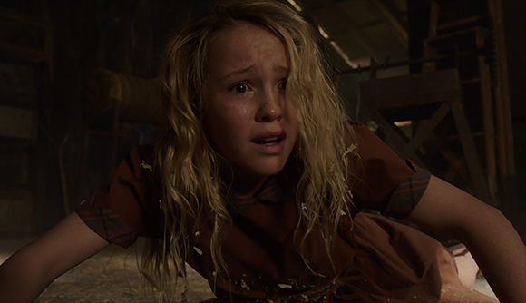 A young blonde girl with a worried expression looking toward the camera in a dimly lit room.