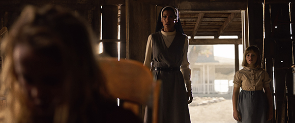 A woman in a grey habit stands in a doorway while a young girl stands to the side in a dimly lit room.
