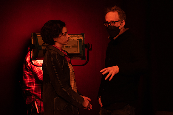 A woman in a dark jacket and a man wearing glasses and a black face mask talk in a dimly lit red room.