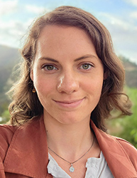 A woman with wavy brown hair and green eyes smiling against a blurred outdoor background.