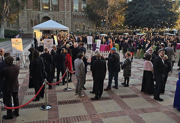 A large group of people dressed formally are gathered outside a building, with red carpet ropes and stanchions guiding them.