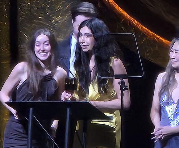 Three women and a man stand behind a podium on a stage. The woman in the center wears a yellow dress and holds a glass. The w