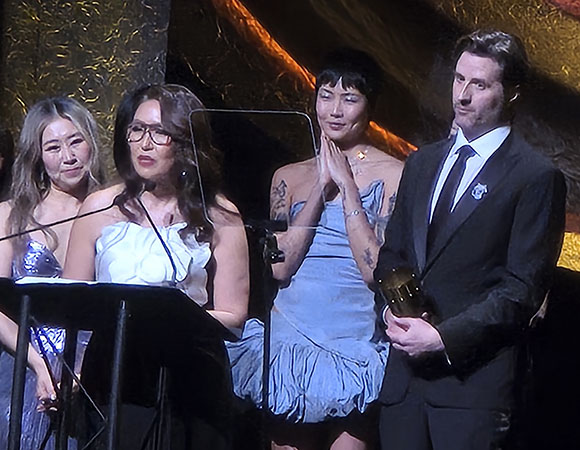 Four people stand on a stage. Two women are at a podium, one speaking. A third person in a blue dress stands behind them with