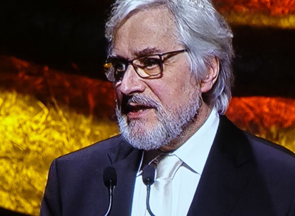 A man with glasses, gray hair, and a beard speaks into microphones.