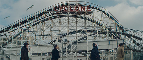 Four people stand in the foreground before the large, wooden Cyclone roller coaster structure under a cloudy sky.
