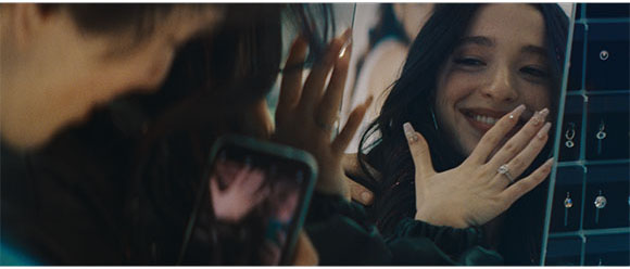 A smiling woman looks at her reflection in a mirror, holding her hands up near her face.