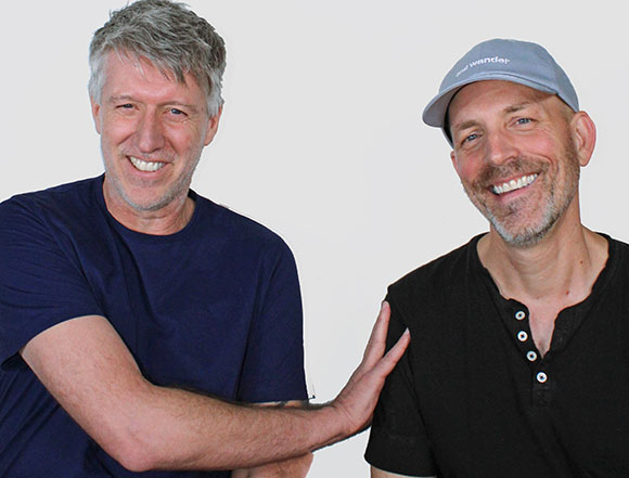 Two smiling men, one in a navy shirt and one in a black shirt and grey cap, against a white background.