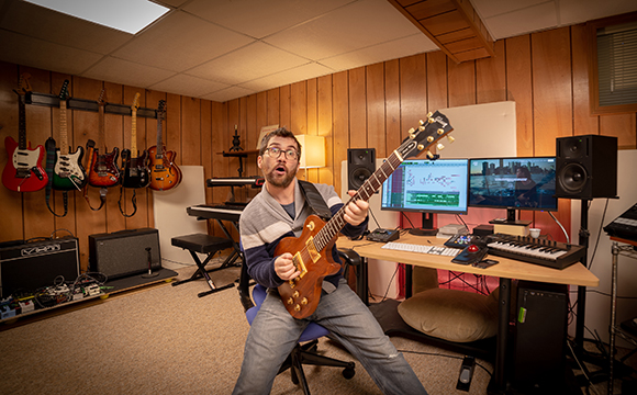 A man sits in a music studio playing a brown electric guitar surrounded by recording equipment.