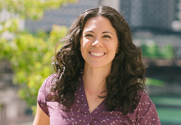 A smiling woman with long, dark curly hair wearing a maroon patterned blouse outdoors.