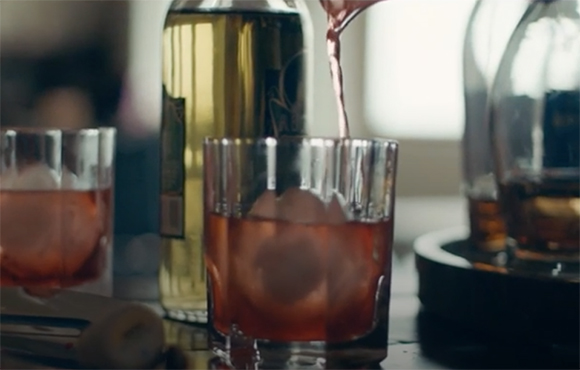 Reddish liquid being poured into a glass containing ice cubes next to a bottle.