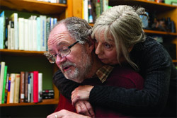 A woman with grey hair leans her head against a man's shoulder as she embraces him from behind.