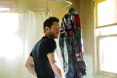 A man in a black t-shirt stands in a room looking at a red and black armored suit hanging on a rack.
