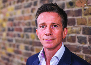A man with short brown hair wearing a white shirt and dark blazer against a brick wall.