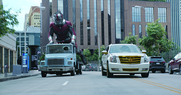 A giant superhero in a red and silver suit stands atop a light blue truck driving down a city street.