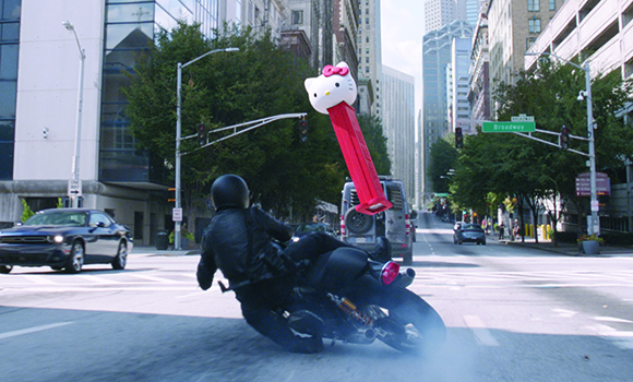 A motorcyclist in black gear leans through a turn on a city street, holding a large pink Hello Kitty prop.