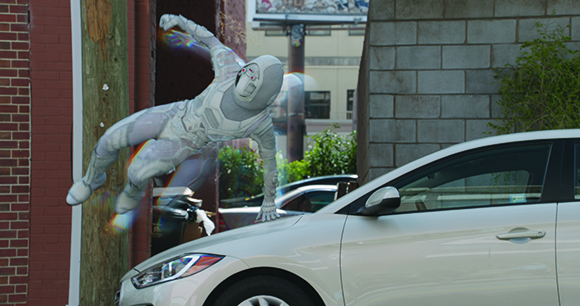 A person in a white, textured bodysuit leaps through the air next to a white car and a brick building.