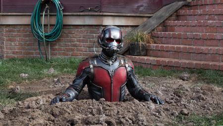 A person in a red and black superhero suit with a helmet emerges from a pile of dirt in a yard.