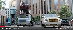 A giant superhero in red and silver armor rides a small blue toy truck down a busy city street.