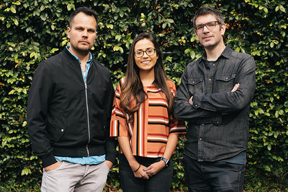 A man in a black jacket, a woman in a striped shirt, and a man in a grey denim shirt stand before a hedge.