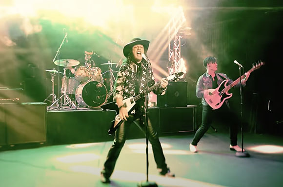A guitarist in a black hat and patterned shirt performs center stage with a bassist and drummer behind him.