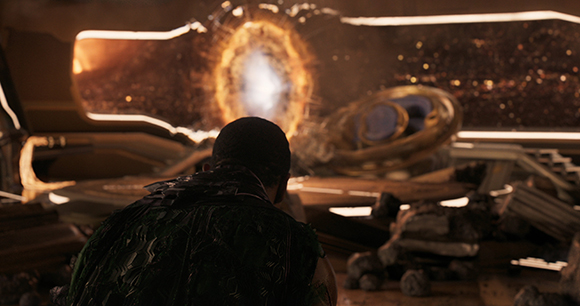 A person in dark armor watches a bright, swirling orange portal in a dimly lit, metallic interior.