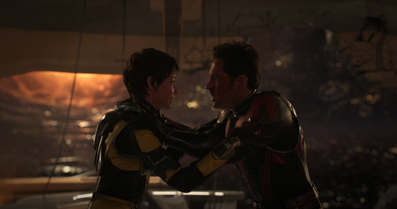 A woman in a yellow and black suit holds the arms of a man in a red and black suit against a dark, glowing background.