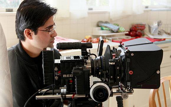 A man with glasses looks through a large professional cinema camera in a kitchen setting.