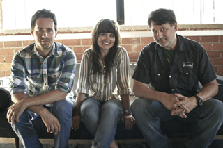 A man in a plaid shirt, a woman in a striped shirt, and a man in a black shirt sitting on a couch.