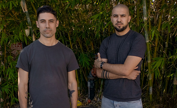 Two men in dark t-shirts standing side-by-side in front of a dense bamboo thicket.