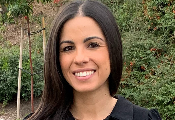 A smiling woman with long dark hair wearing a black shirt, standing in front of green bushes.