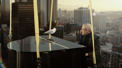 A man sits at a black grand piano suspended by yellow straps over a cityscape at sunset.