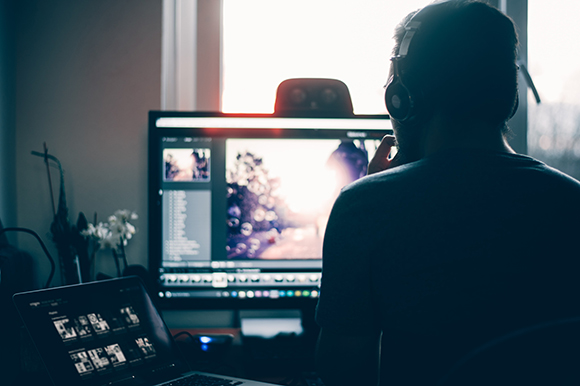 A person in silhouette wearing headphones looks at a large computer monitor displaying photo editing software.