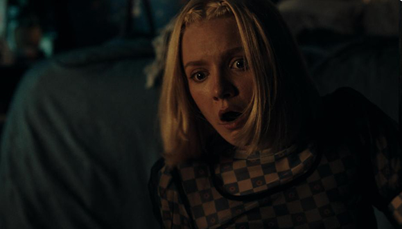 A blonde woman looks toward the camera with wide eyes and an open mouth in a dark setting.
