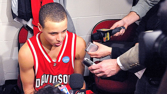 A basketball player in a red jersey speaks into several microphones held by reporters.