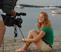 A smiling woman with long red hair sits on a sandy beach while a camera operator films her.
