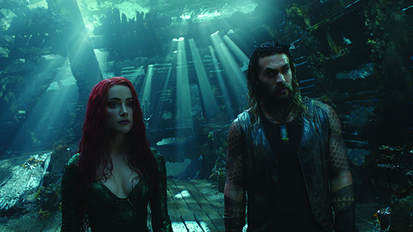 A man with dark hair and a woman with long red hair stand in a sunlit underwater ruins.