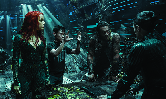 A director gestures toward actors on a dark, textured film set designed to look like an underwater ruin.