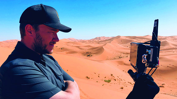 A man in a black cap and shirt looks at a camera monitor in a vast orange sand dune desert.