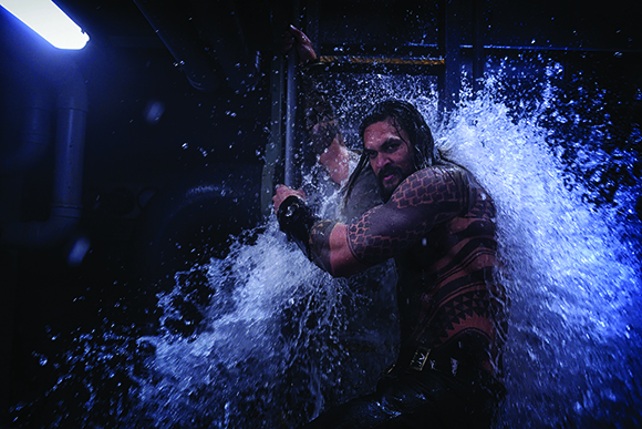 A tattooed man is partially obscured by a large, dramatic splash of water in a dark setting.