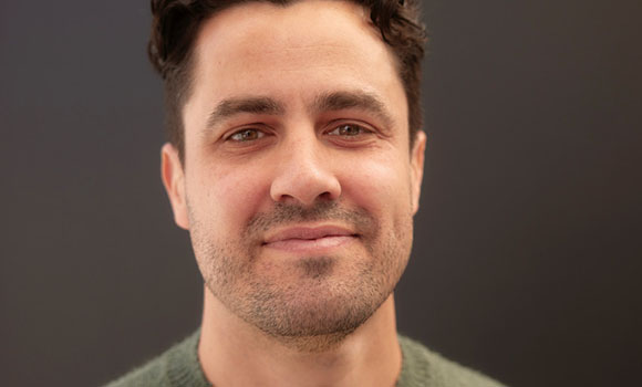 A man with dark curly hair and light stubble smiles against a dark gray background.