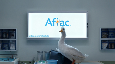 A white duck stands in a medical setting in front of a bright Aflac logo on a screen.