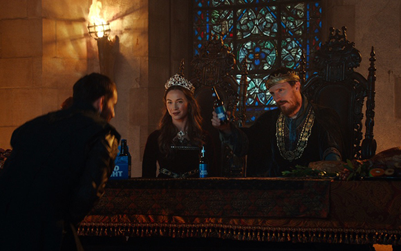 A man and woman in royal medieval clothing sit at a long table in a stone castle hall.