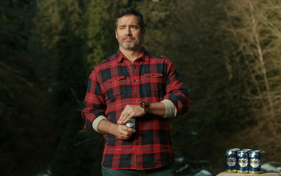 A man in a red and blue plaid shirt holds a blue beer can in a wooded outdoor setting.