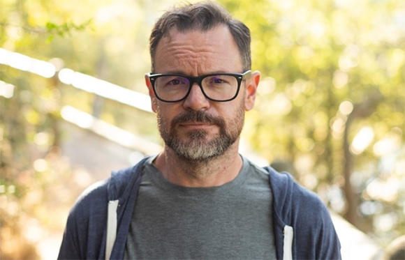 A man with short dark hair, glasses, and a salt-and-pepper beard looking forward outdoors.