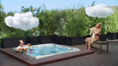 A man relaxes in a hot tub while a woman sits on a bench holding a small white dog on a wooden deck.