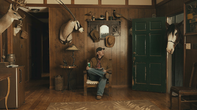 A man in a cap and jacket sits in a wooden chair holding a drink in a room filled with mounted animal heads.
